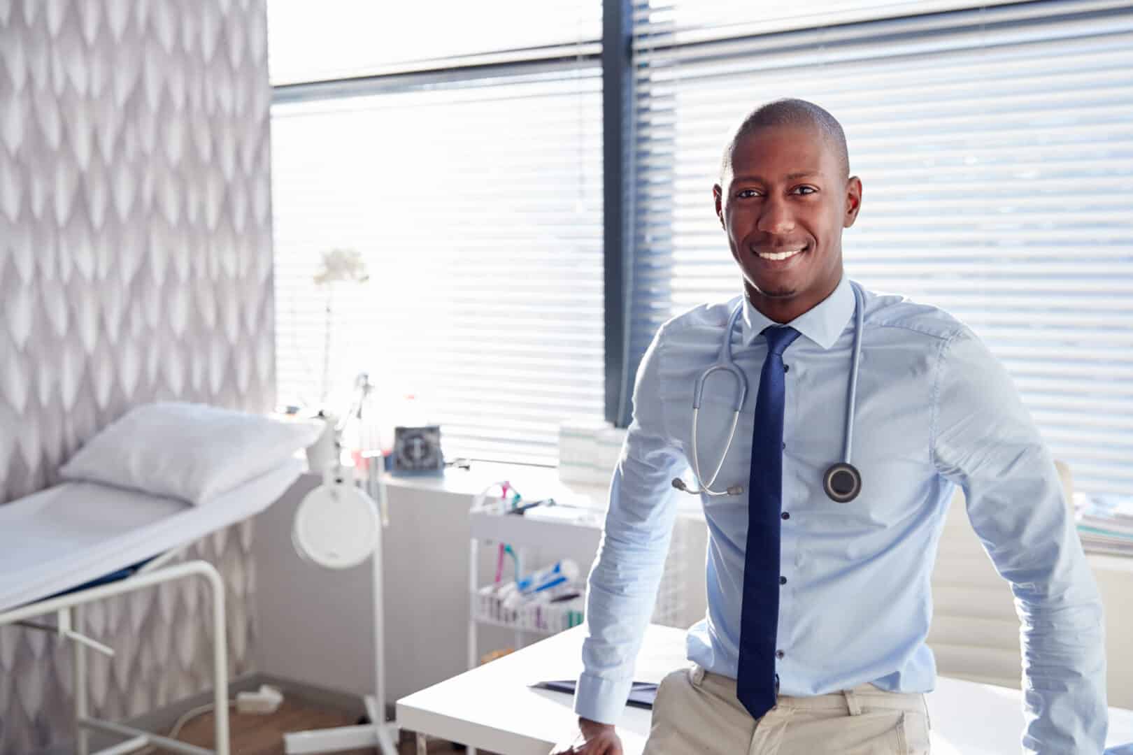 A doctor wearing a stethoscope around his neck, leaning on an examination table in an examination room.